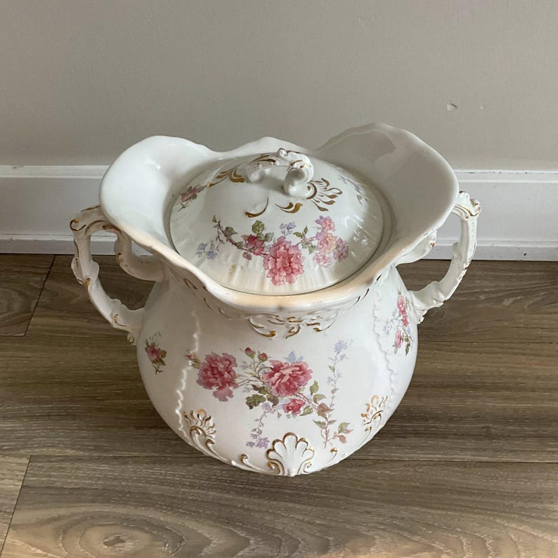 May include: A white porcelain chamber pot with a lid, adorned with pink floral patterns and gold accents. The pot features two handles and a scalloped rim, showcasing a vintage aesthetic.