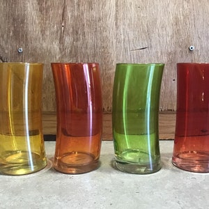May include: Four glass tumblers in different colors: yellow, orange, green, and red. The glasses are curved and have a clear, smooth finish.