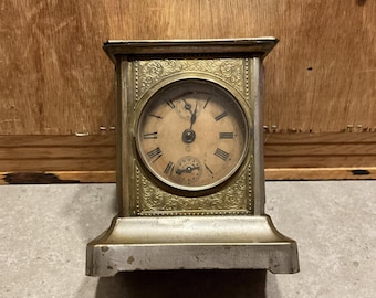 Könnte beinhalten: Eine antike silber- und goldfarbene Uhr mit rundem Zifferblatt und römischen Ziffern. Die Uhr hat ein verziertes, graviertes Design um das Zifferblatt und ein kleineres Zifferblatt darunter. Die Uhr steht auf einem rechteckigen Sockel.