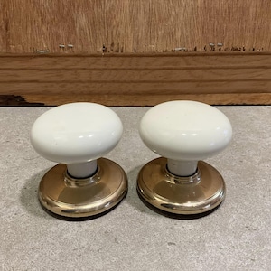 May include: Two white porcelain door knobs with brass bases. The knobs are round and have a smooth finish.