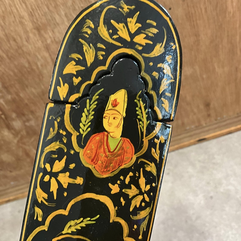 May include: A black, hand-painted wooden box with a hinged lid. The box features a detailed design with gold floral patterns and a central portrait of a person in traditional attire. The person is wearing a yellow hat and an orange robe.