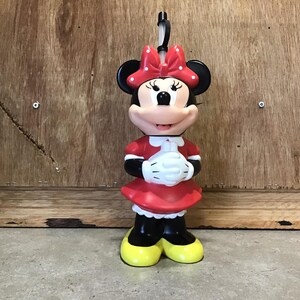 May include: A Minnie Mouse figurine, featuring a red dress, white gloves, and a polka-dot bow. The figure has black ears, yellow shoes, and a smiling expression. A black loop is attached to the top of the head.