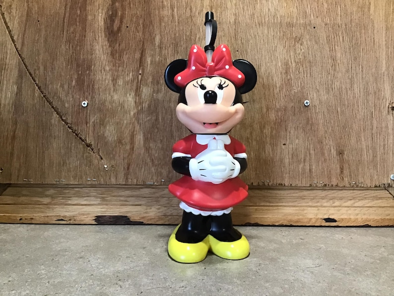 May include: A Minnie Mouse figurine, featuring a red dress, white gloves, and a polka-dot bow. The figure has black ears, yellow shoes, and a smiling expression. A black loop is attached to the top of the head.
