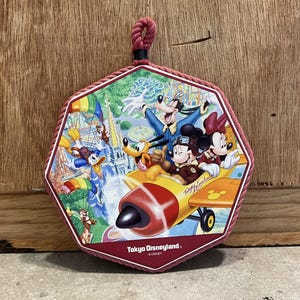 May include: A red octagonal-shaped wall hanging with a rope loop at the top. The wall hanging features a colorful illustration of Mickey Mouse, Minnie Mouse, Goofy, and Pluto flying in a red and yellow airplane. The text "Tokyo Disneyland" and "© DISNEY" are printed on the wall hanging.