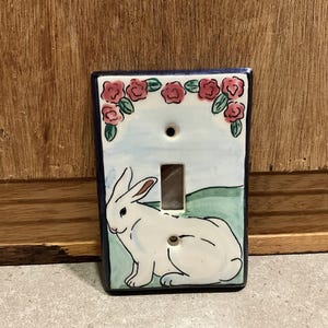 May include: Hand-painted ceramic light switch cover featuring a white rabbit and pink roses. The rectangular cover has a blue border and a toggle switch opening. The background includes a light blue sky and green hills.