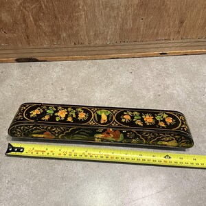 May include: A black, rectangular box with a hinged lid, adorned with floral and figural designs in yellow, green, and gold. The box is approximately 14 inches long. A yellow measuring tape is placed next to the box.
