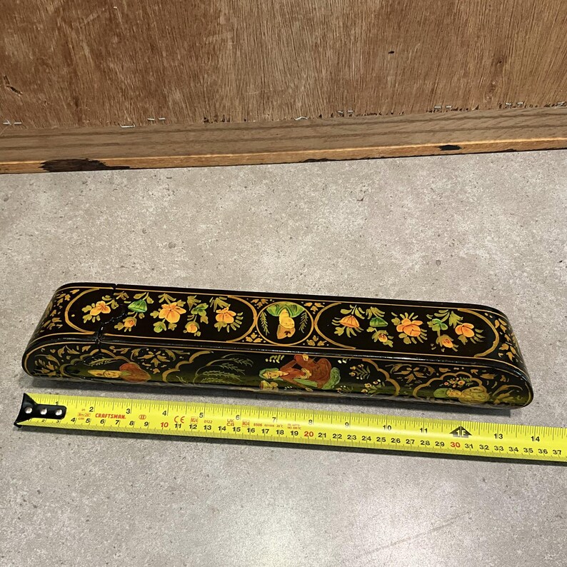 May include: A black, rectangular box with a hinged lid, adorned with floral and figural designs in yellow, green, and gold. The box is approximately 14 inches long. A yellow measuring tape is placed next to the box.