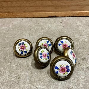 Op de afbeelding: Zes vintage kastknoppen met een bloemenontwerp. Elke knop heeft een wit keramisch oppervlak met een roze en blauw bloemmotief, gezet in een messingkleurig metalen frame. De knoppen zijn rond en hebben een klassieke, decoratieve stijl.