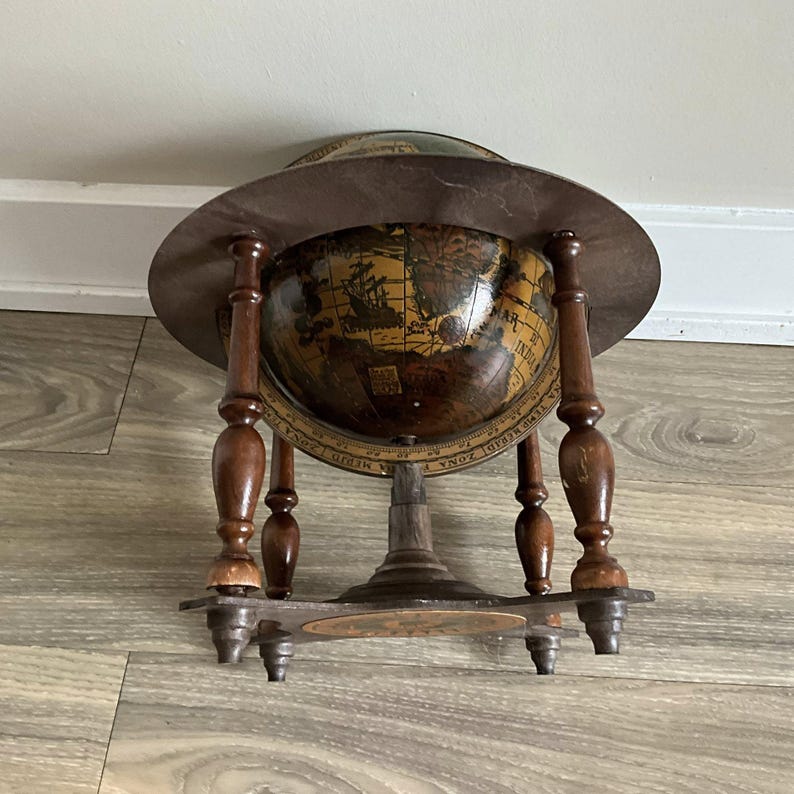May include: A vintage globe with a dark brown wooden stand and a circular, aged metal shade. The globe itself features a detailed map with sepia tones and nautical illustrations. The stand has four ornate legs and a lower shelf.
