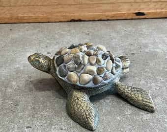 May include: A decorative turtle figurine with a shell made of small, round stones in shades of brown, gray, and white. The turtle is made of a metallic material with a green and gold finish.