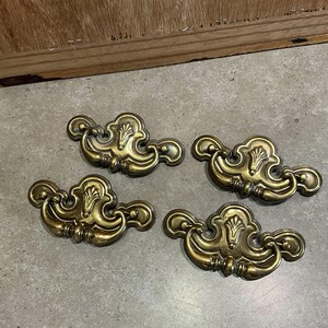 May include: Four ornate brass cabinet pulls with a decorative floral design. Each pull has a curved shape with a central loop for pulling.