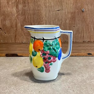 May include: A white ceramic pitcher with a blue rim and handle. The pitcher is decorated with a colorful fruit design featuring oranges, pears, grapes, and leaves.