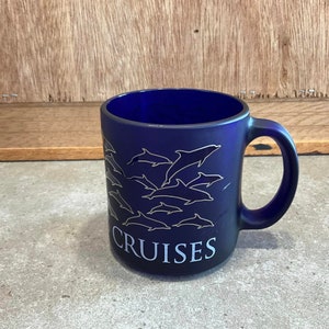 May include: A cobalt blue ceramic mug featuring a silver dolphin design and the word "CRUISES" printed on the front.