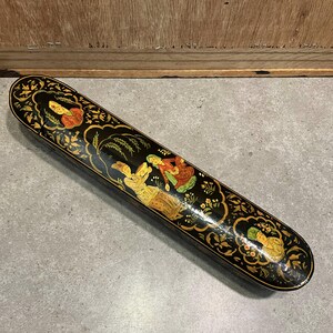May include: A long, black decorative box with a detailed, hand-painted design. The box features gold floral accents and illustrations of figures in colorful clothing. The box is likely made of wood and has a glossy finish.