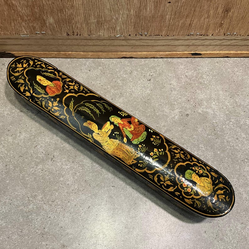 May include: A long, black decorative box with a detailed, hand-painted design. The box features gold floral accents and illustrations of figures in colorful clothing. The box is likely made of wood and has a glossy finish.