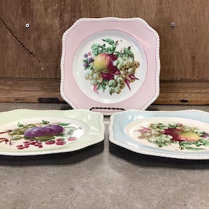 Puede incluir: Tres platos cuadrados de cerámica con bordes festoneados. Cada plato presenta un fondo de color diferente y un diseño central de frutas y uvas. Los platos están dispuestos en fila sobre una superficie de madera.