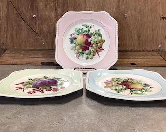 Puede incluir: Tres platos cuadrados de cerámica con bordes festoneados. Cada plato presenta un fondo de color diferente y un diseño central de frutas y uvas. Los platos están dispuestos en fila sobre una superficie de madera.