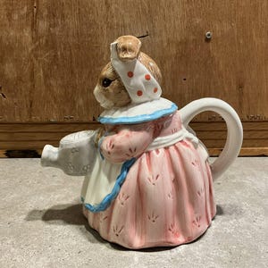 Może przedstawiać: Ceramiczny czajnik w kształcie królika w czepku i sukience. Czajnik jest głównie różowy i biały z niebieskimi akcentami. Królik trzyma dzióbek konewki. Uchwyt ma zakrzywioną białą konstrukcję.
