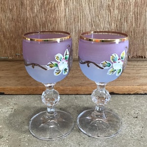 May include: Two purple glass wine glasses with gold rims and painted floral designs. The glasses have a clear stem and base.