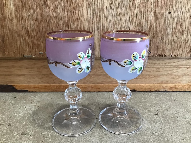 May include: Two purple glass wine glasses with gold rims and painted floral designs. The glasses have a clear stem and base.