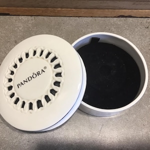 May include: White Pandora jewellery box with an open lid. The lid has a decorative, openwork design around the edge and the Pandora logo in the centre. The interior of the box is lined with black velvet.