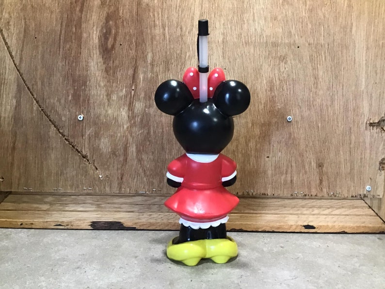 May include: A Minnie Mouse-shaped tumbler with a red dress, white trim, and yellow shoes. The black head features large ears and a red bow with white polka dots. A black and white straw extends from the top.