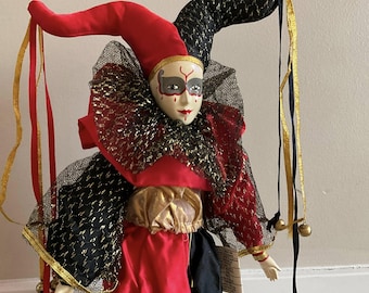 Op de afbeelding: Een porseleinen narrenpop met een rood-zwart kostuum, een zwart en gouden net en een zwart masker met witte en rode details. De pop heeft een goud-rode hoed met een lange zwart-gouden streamer.