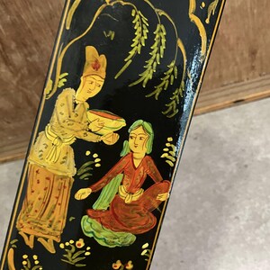 May include: Close-up of a black, hand-painted decorative panel featuring two figures in traditional attire. One figure is offering a bowl to another seated figure. The artwork is in shades of yellow, red, and green, with gold accents and floral details.