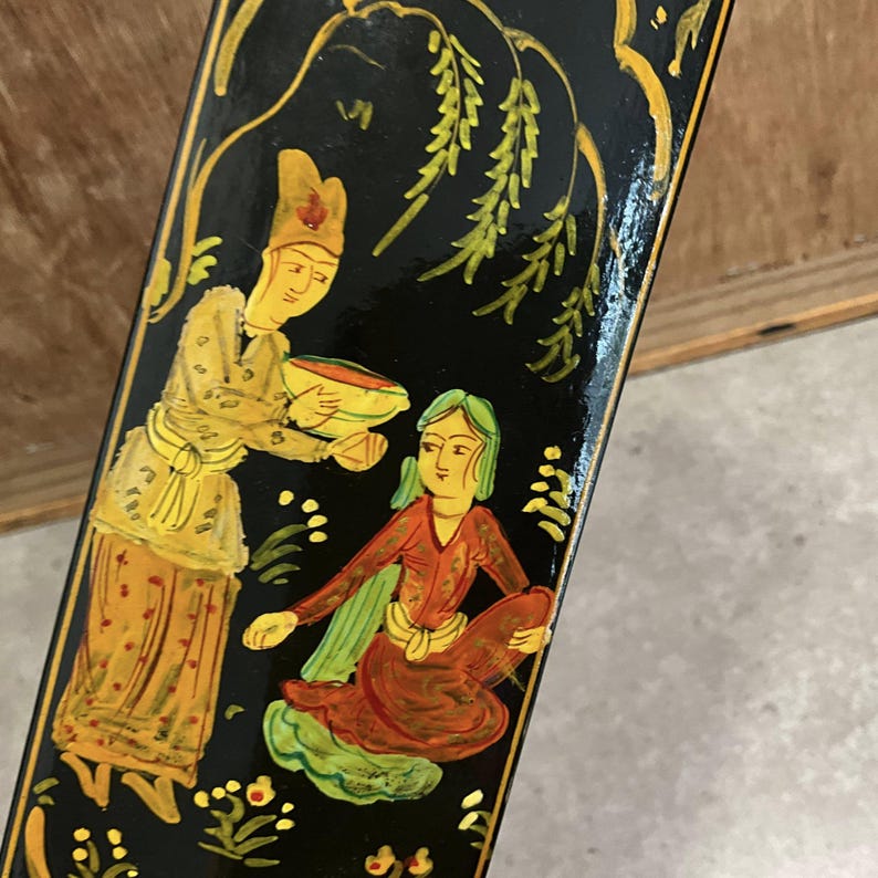 May include: Close-up of a black, hand-painted decorative panel featuring two figures in traditional attire. One figure is offering a bowl to another seated figure. The artwork is in shades of yellow, red, and green, with gold accents and floral details.