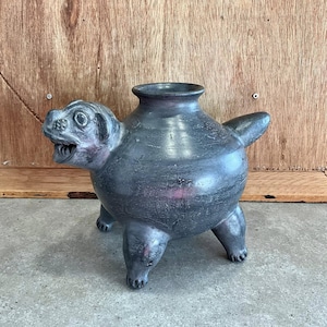 Pode incluir: Um vaso cinza escuro em cerâmica em forma de cão, com um corpo largo, uma borda alargada e três pernas. A cabeça do cão é detalhada com boca aberta e orelhas. O vaso tem cerca de 20 cm de altura.