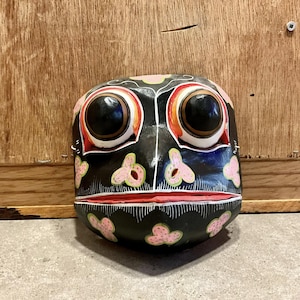 May include: A black wooden mask with pink and green floral designs, large black eyes, and a red mouth.