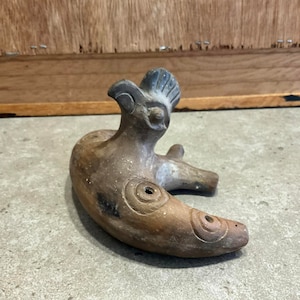 May include: A brown ceramic whistle shaped like a bird with a crested head. The whistle has a crescent-shaped body with circular designs and holes.