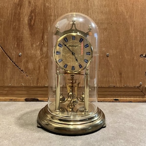 May include: A brass-colored antique clock with a glass dome. The clock face is gold with blue Roman numerals. The pendulum and base are also brass. The clock is a decorative item.