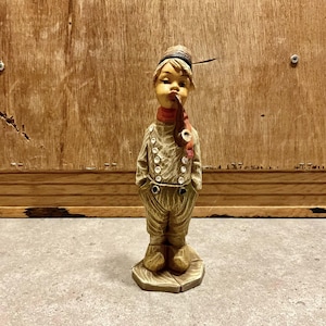 Puede incluir: Figura vintage de un joven con pipa. La figura lleva un sombrero, una bufanda roja y un atuendo abotonado. La figura es de color beige y marrón, con las manos en los bolsillos. La figura está sobre una pequeña base beige.