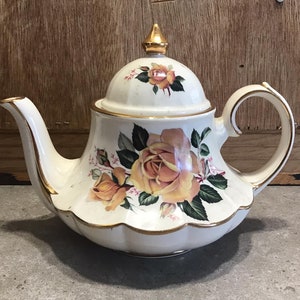 Könnte beinhalten: Eine weiße Teekanne mit goldener Verzierung und einem Blumendesign mit gelben Rosen. Die Teekanne hat einen Deckel mit einem goldenen Knopf.