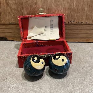 Peut inclure: Deux boules Yin Yang bleues et blanches avec une finition bleu canard foncé, devant une boîte rouge ouverte avec un fermoir doré. La boîte a un intérieur en velours rouge et un petit morceau de papier à l'intérieur.