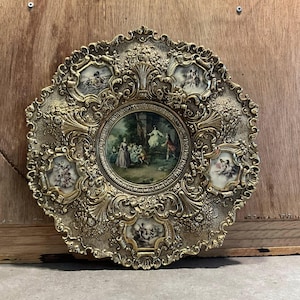 Pode incluir: Prato decorativo dourado ornamentado com uma imagem central circular de um grupo de pessoas num cenário florestal. O prato apresenta um design detalhado e com rolagem e seis imagens menores. Artigo vintage.