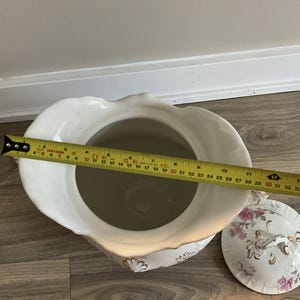 May include: A white porcelain chamber pot with a lid, featuring floral and gold accents. The pot measures approximately 10 inches in diameter. The lid has a handle and a floral design. The pot is on a wooden surface.