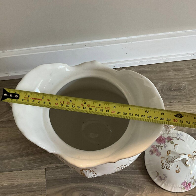 May include: A white porcelain chamber pot with a lid, featuring floral and gold accents. The pot measures approximately 10 inches in diameter. The lid has a handle and a floral design. The pot is on a wooden surface.