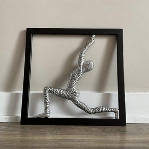 Op de afbeelding: Een zilveren metalen sculptuur van een persoon in een yogapose, ingelijst in een zwart frame.