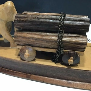May include: A wooden elephant figurine pulling a cart with two logs secured by a chain. The cart has two wooden wheels and sits on a wooden base.