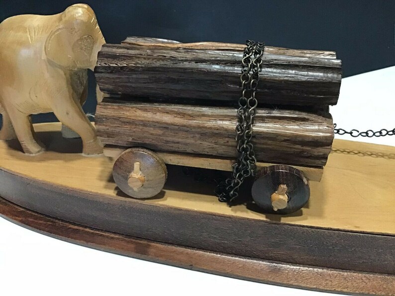 May include: A wooden elephant figurine pulling a cart with two logs secured by a chain. The cart has two wooden wheels and sits on a wooden base.