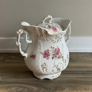 May include: White porcelain pitcher with a lid, adorned with pink floral patterns and gold accents. The pitcher features ornate handles and a decorative spout. A vintage-style decorative piece.