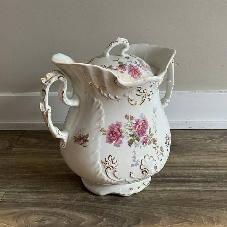 May include: White porcelain pitcher with a lid, adorned with pink floral patterns and gold accents. The pitcher features ornate handles and a decorative spout. A vintage-style decorative piece.