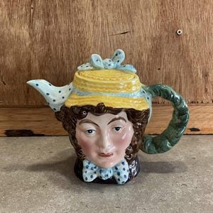 May include: A ceramic teapot shaped like a woman's head. The teapot is decorated with blue and white polka dots, a yellow straw hat, and a green handle.