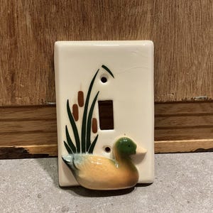 Peut inclure: Plaque d'interrupteur en céramique crème avec un motif de canard et de quenouilles. Le canard a une tête verte, un corps orange et un bec blanc. Les quenouilles sont marron et vertes. La plaque a une ouverture rectangulaire.