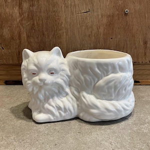 May include: A white ceramic cat planter with red eyes. The planter is shaped like a fluffy cat lying down, with a round opening for plants. The cat has detailed fur and a whimsical design, perfect for a cat lover's home decor.