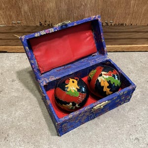May include: A blue decorative box with a red interior holds two black Chinese health balls. One ball features a colorful bird design, and the other has a swirling pattern in red, green, and yellow. The box has a gold clasp.