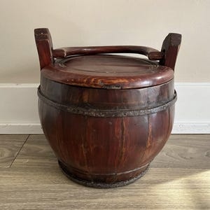 May include: A dark brown wooden bucket with a lid and a handle. The bucket has a rounded shape with metal bands around the middle and base. The wood grain is visible, and the bucket appears to be well-worn. A vintage item.