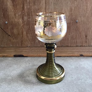 Könnte beinhalten: Ein grünes Glasgoblet mit goldfarbenem Rand und einem gravierten Traubenmuster. Das Goblet hat eine breite Schale und einen hohen, schlanken Stiel.